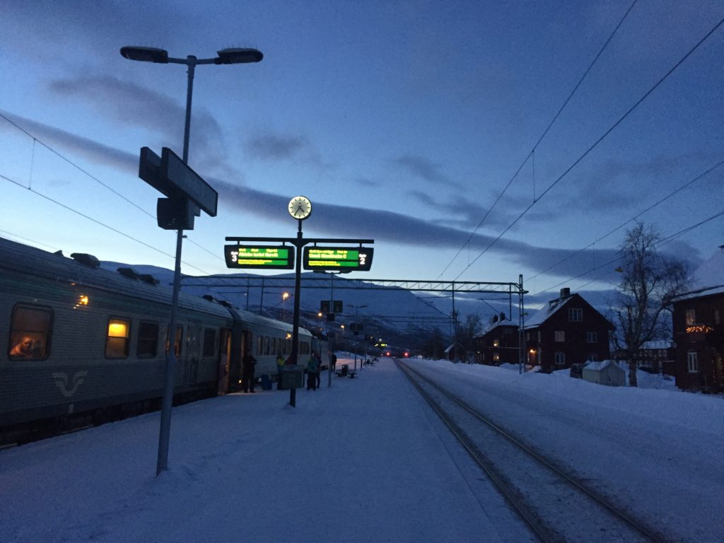 Abisko Ostra train station in Swedish Lapland, with brilliant blue hour lighting, one train, and a few houses visible in the area, mountains behind the train.