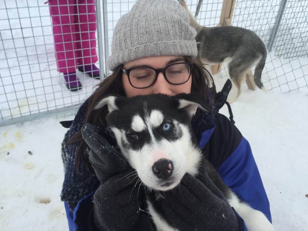 Allison in glasses and a grey hat, with her eyes closed, smiling as she cuddles a black and white husky puppy with one brown eye and one blue eye.