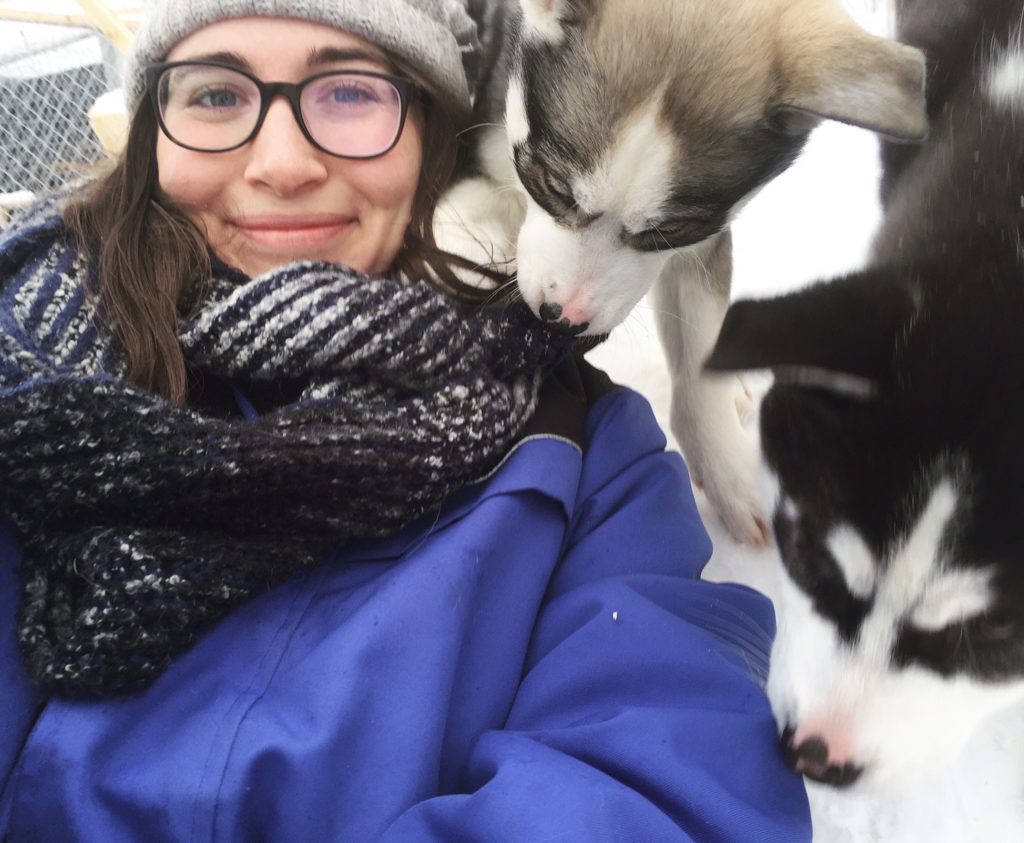 Allison smiling while wearing a hat, scarf, glasses, and a blue thermal suit, with two huskies pawing at her playfully.