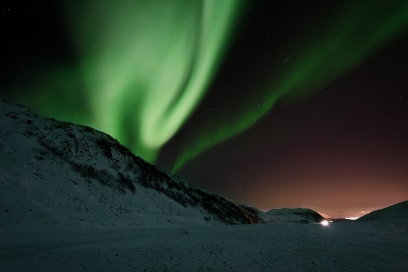 The green waves of the northern lights as seen from above a mountain in sweden with lights on the horizon