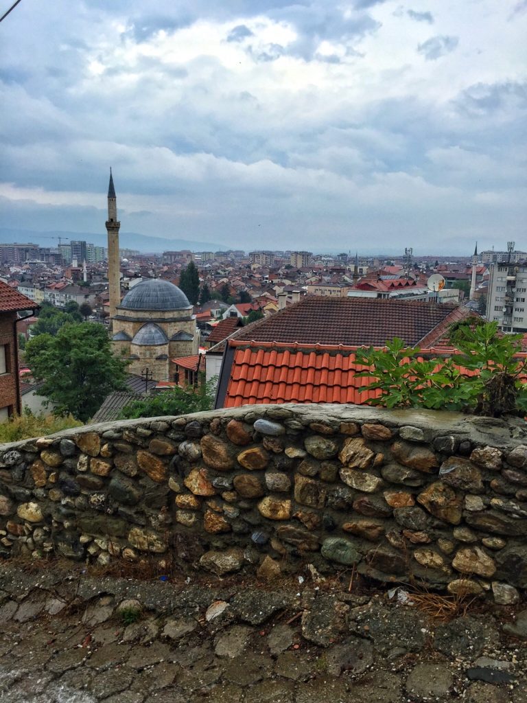 The gorgeous main mosque in Prizren, Kosovo, with a stone wall in the foreground and tiled roofs of nearby buildings.