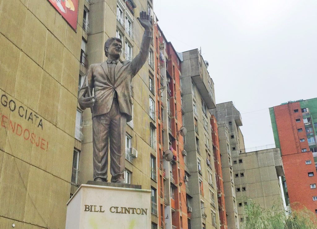 The statue of Bill Clinton with very large hands and buildings of downtown Pristina behind the bronze statue