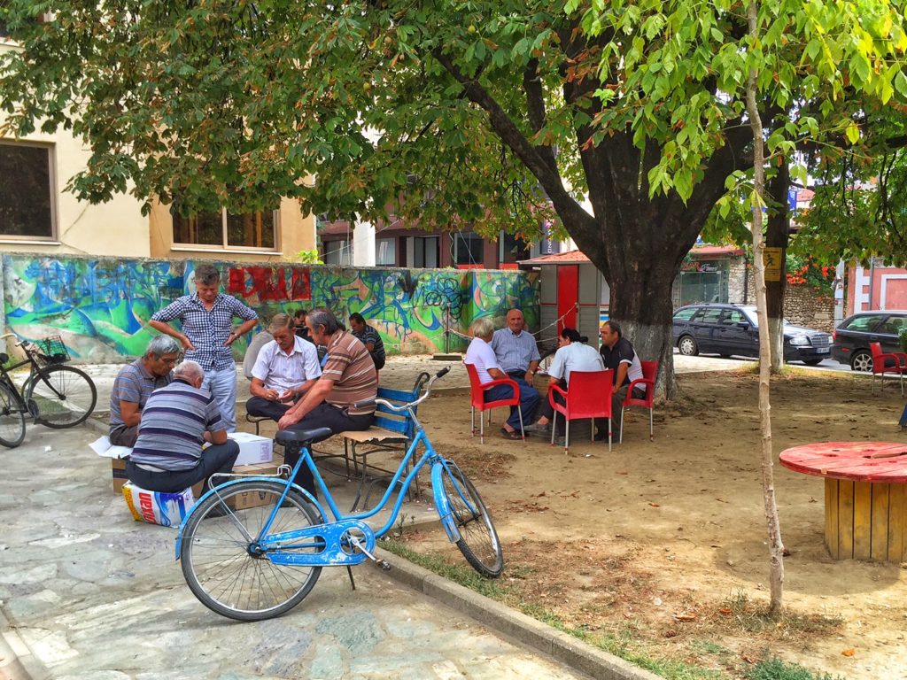 Men sitting around on boxes and chairs playing cards in a public park, with a blue bike and a black bike 