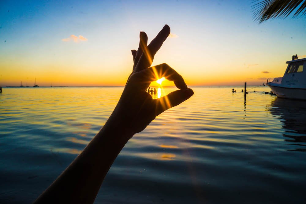 Allison giving an OK signal like scuba divers do while looking at the sunset, with a sunburst, on Caye Caulker