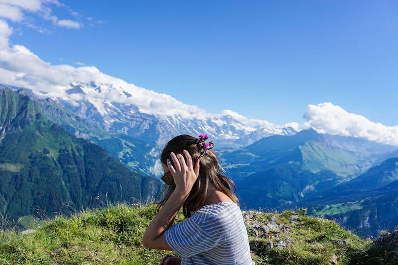 allison with flowers in her hair, head tilted down, on the top of the mountains with snow-capped peaks visible covered in clouds in the distance
