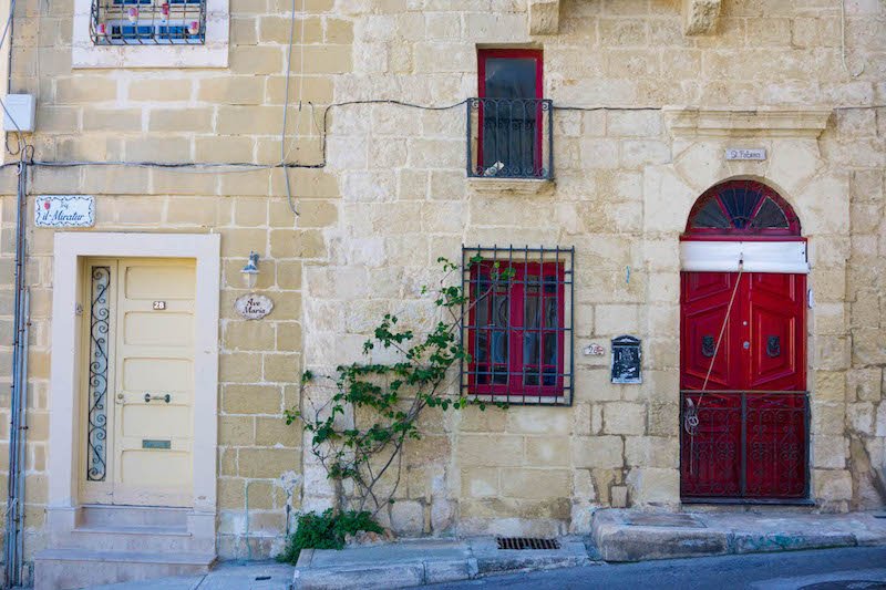 A red door and a red window against sandstone bricks on a historic building in Birgu