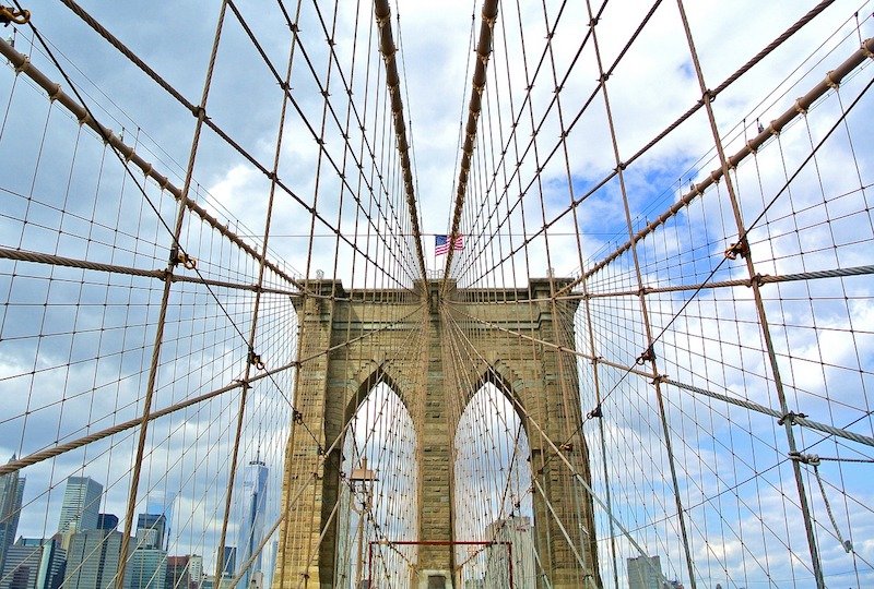 view of the beautiful symmetry of the suspension bridge of the brooklyn bridge with an american flag on top and the new york skyline in the background