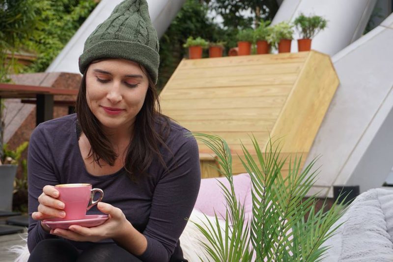 Allison Green enjoying a day in Taiwan with a pink cup of coffee in her hand, smiling while wearing a green hat and purple long sleeve shirt in winter in Taiwan.