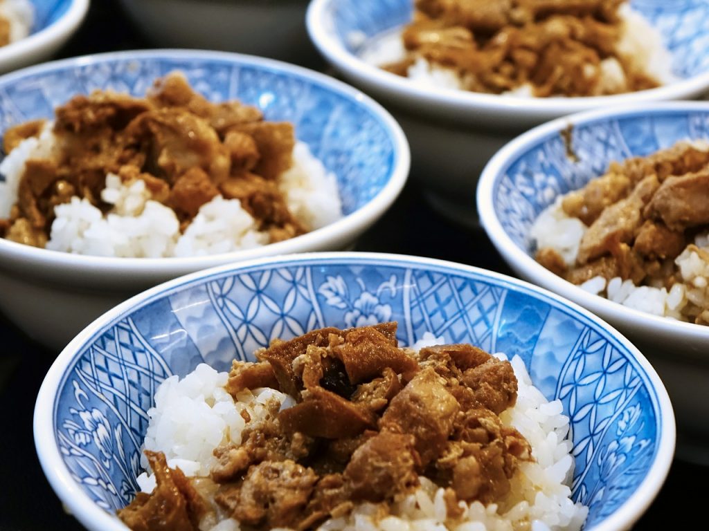 a bowl of pork braised rice, with pork belly served on top of white rice in a blue and white bowl. three bowls visible in the frame.