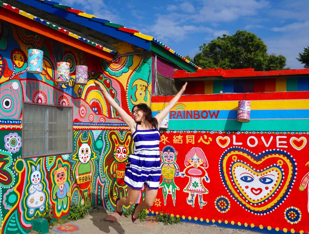 Allison Green jumping with white and blue dress in a colorful part of a rainbow village where it says rainbow and love