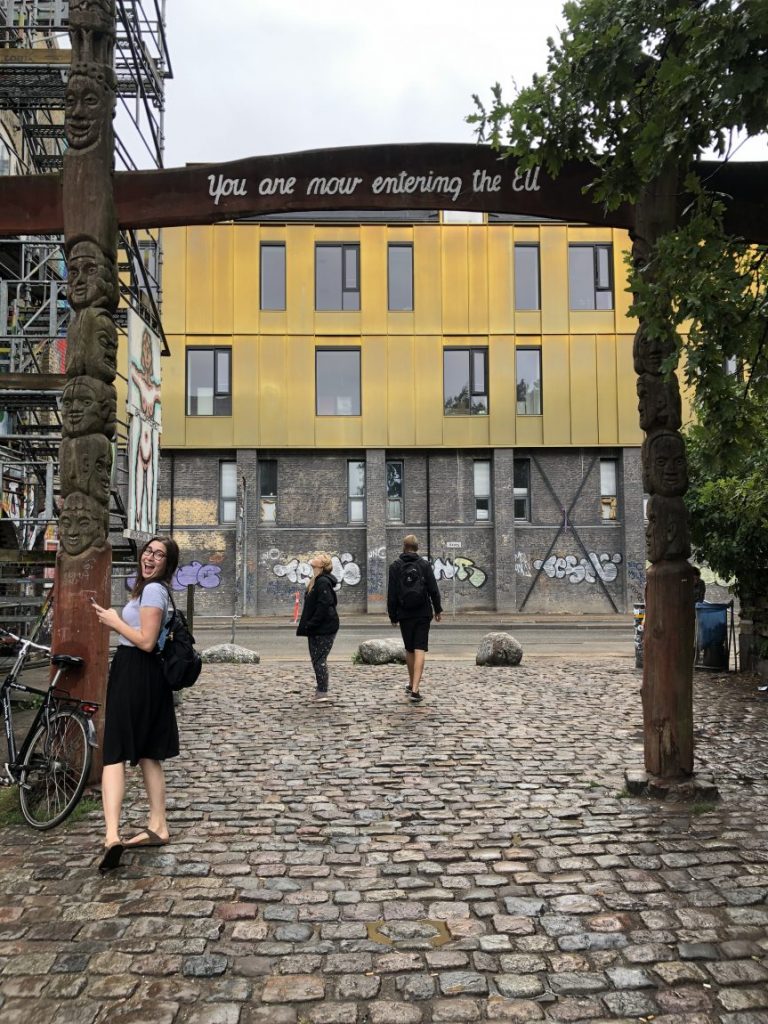 Allison Green smiling as she enters Freetown Christiania in copenhagen with a white shirt and black skirt and backpack