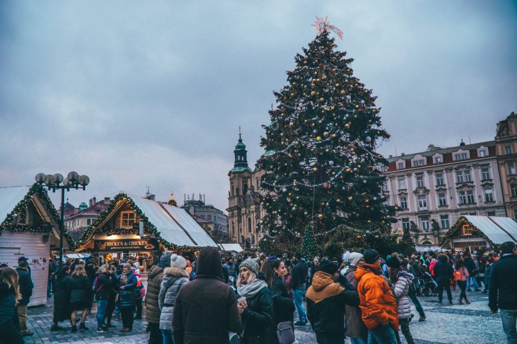 A bunch of visitors and locals alike enjoying the festive atmosphere of the Prague Christmas market on an overcast day in December. You can see a large Christmas tree in the Old Town Square and several wooden stalls adorned in lights and evergreen brush foliage.