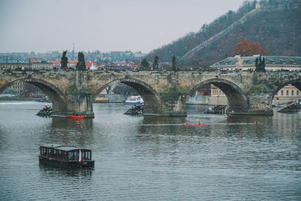 Snow falling on the Vltava River of Prague with a clear view of the Prague Bridge in the center, below there are people in a red kayak on the river. You can see people walking on the bridge and the famous statues on the bridge as well.