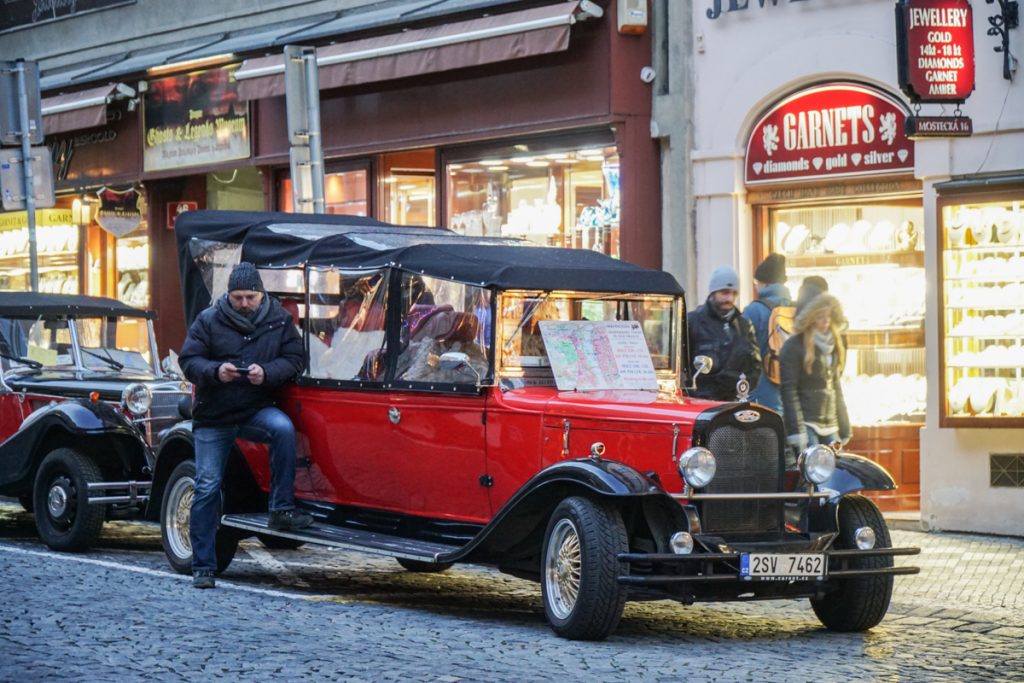 Man on his phone in Prague in winter waiting for customers to approach him for a classic car tour of Prague, leaning on a red car with a temporary roof