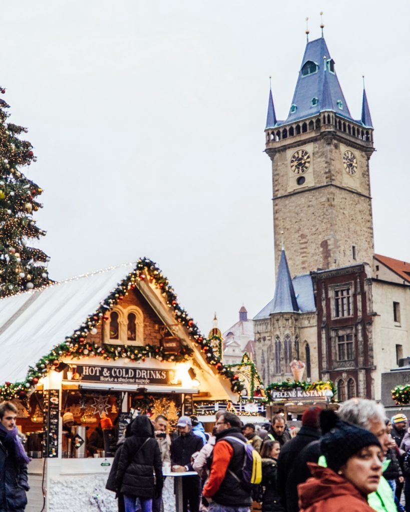 Close-up view of fellow travelers in the Old Town Christmas market area with a giant astronomical clock in the background, and foreground shows the Christmas market stalls and Christmas tree in the square.