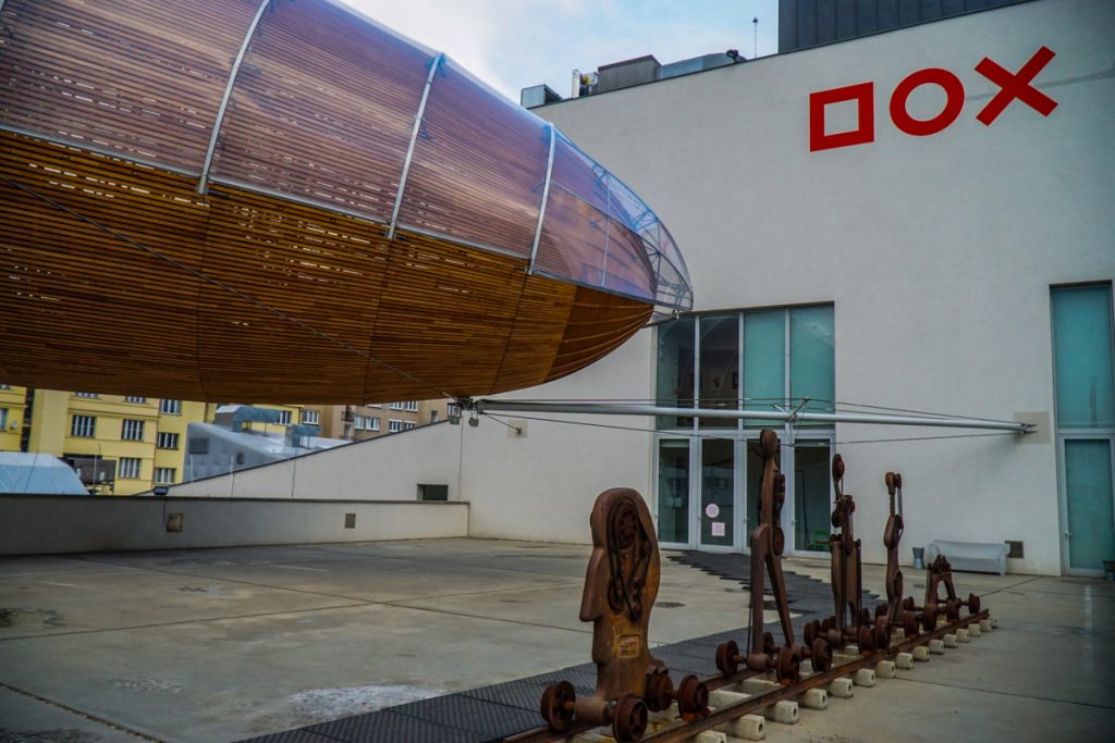 View of a blimp-like massive sculpture on the roof of the DOX museum, made of wood, as well as other contemporary sculptures on display in a modern art museum in Prague.