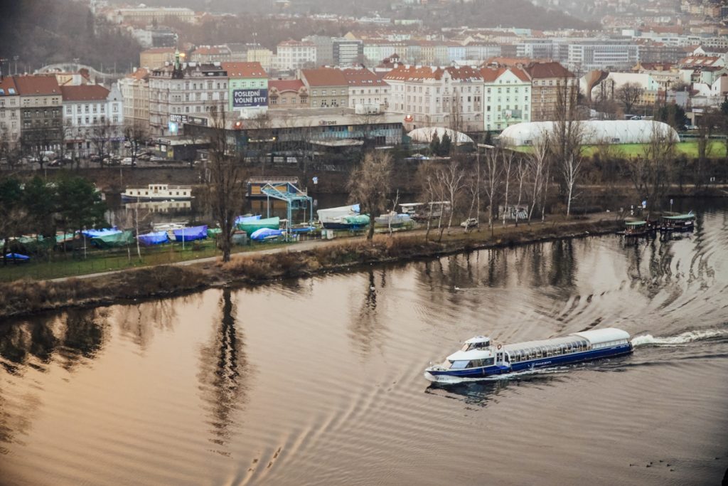 View from above of a boat passing along the Vltava River, with view of the Czech cityscape behind the river, as the sun sets in winter