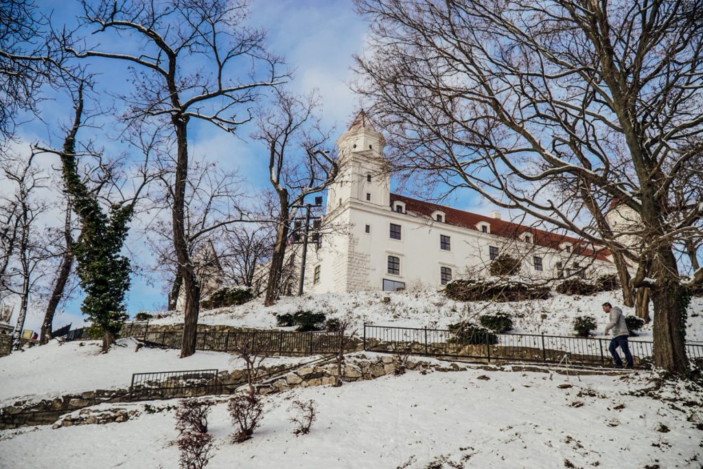 The all-white Bratislava Castle as seen from lower on the hill, covered in snow as you ascend the steps towards the castle, with barren trees around.