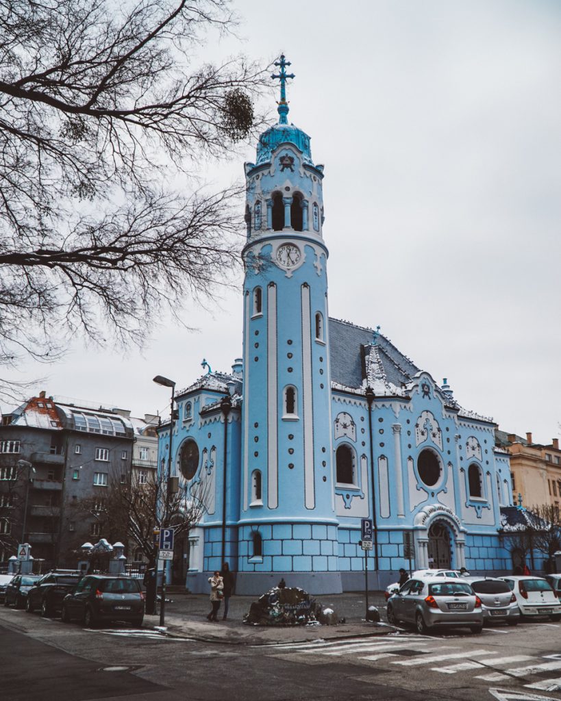 A blue church in the winter with snow on the ground with a cloudy sky in the background.