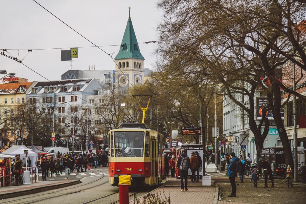 A winter scene in Bratislava, with a tram running through the center, a church in the background, and lots of people shopping at outdoor Christmas markets on a popular street.