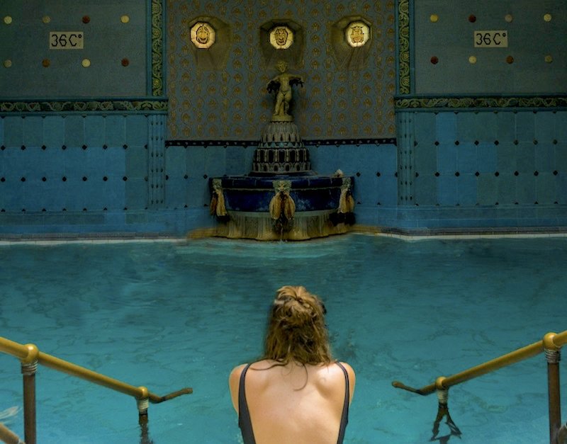 Allison Green, wearing a backless bathing suit in black, with her hair half-pinned-up, sitting in front of the 36 degree indoor thermal bath in Gellert Baths in Budapest, a beautiful indoor bathhouse in an Art Nouveau style, with lots of blue tile.