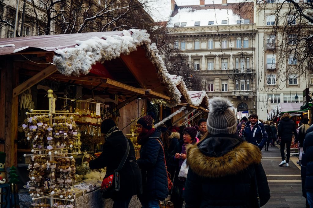 The back of someone’s head with a puffball hat, walking through the Christmas markets of Budapest in December, looking at wooden souvenirs and street stalls in the market. 