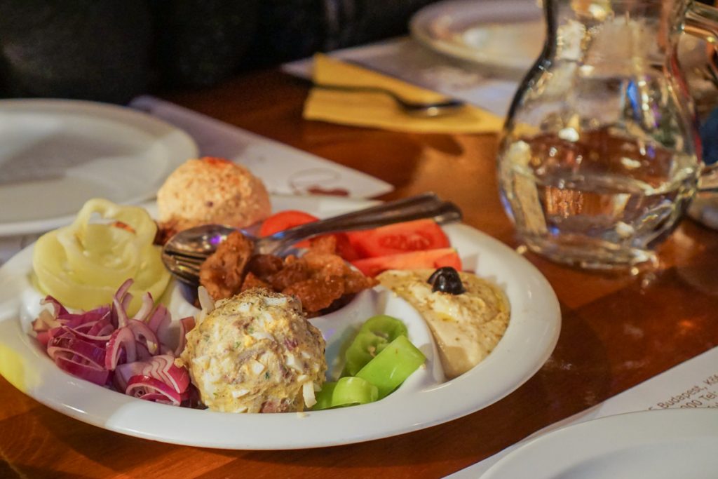 A delicious plate of food in a Budapest restaurant with red onion, a ball of pate and other things like chopped hard boiled egg, hummus, cracklings, and a glass of wine.