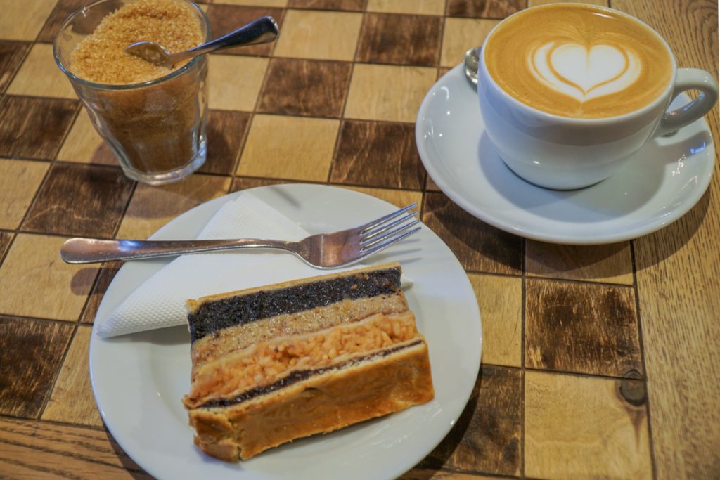 A layered cake with different flavors like nuts and poppy seeds, next to a cup of coffee with beautiful latte art, on a checkered table in a cafe in Budapest.