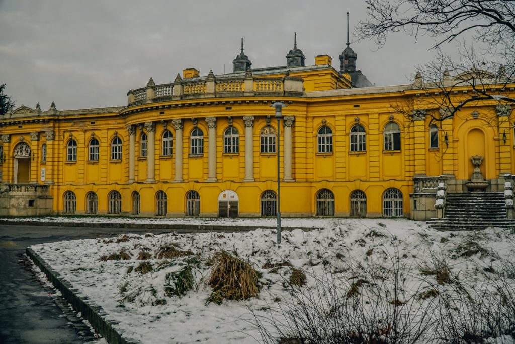 A bright yellow building in Budapest, which stands out brightly against the gray sky and the white snowy landscape. This is a thermal bath house located in a park.