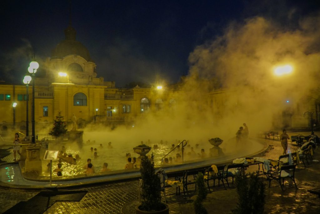 A view of the Szechenyi baths outdoor area in the pitch black night in the middle of winter in Budapest, when the thermal baths are a great place tow arm up. Neoclassical architecture and large outdoor bath with steam rising.