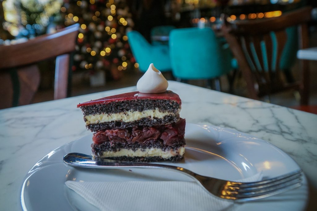 A slice of chocolate cake with layers of raspberry interior and also a creme interior with a dollop of cream atop. You can see the details of a Christmas tree in the background inside a fancy cafe.