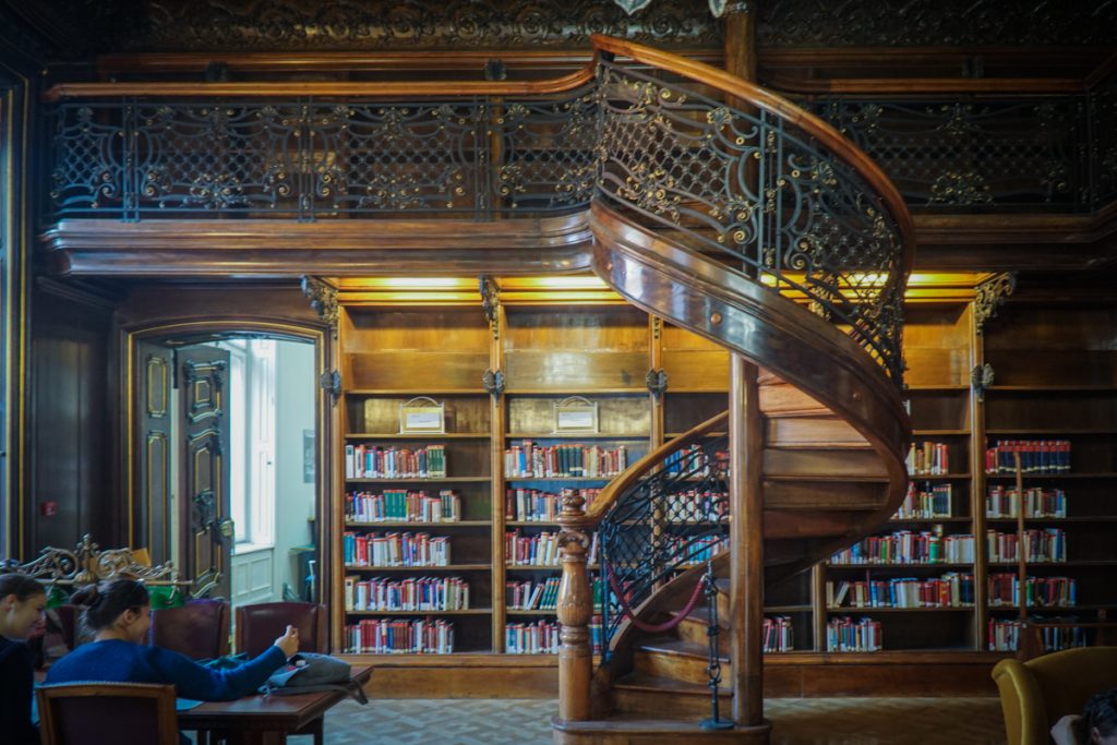 A beautiful spiral wood staircase, with library details and books and people enjoying the reading room in a pretty Budapest library.