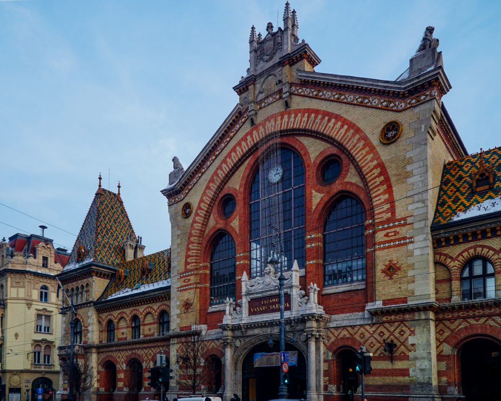 
The exterior of the Budapest Market Hall, with a little dusting of snow on the colorful mosaic roof, with beautiful blue sky behind it on a wintery day.