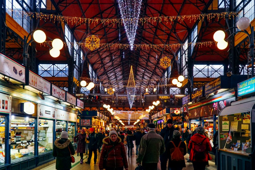 
The interior view of the Budapest Market Hall in winter, with people in winter coats enjoying a break from the cold, with a festively lit and decorated food hall.