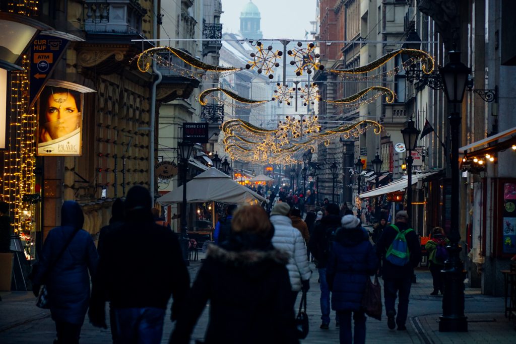 People in their winter outfits, wearing winter jackets and hats, etc. strolling down the festively-lit street called Vaci Utca which is one of the most popular pedestrian paths in the city.