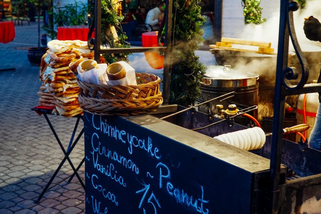 A stall in Budapest Christmas market selling Chimney Cakes with cinnamon vanilla and cocoa flavors, as well as other baked goods, in the evening light.