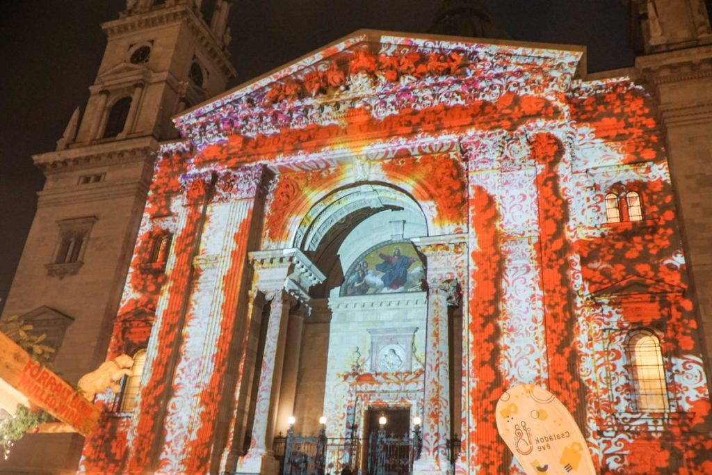 Facade of the St Stephen Basilica in Budapest, a beautiful large church. The facade has a bright red and floral patterned visual projection on the building face, creating a beautiful Christmas-themed light show.