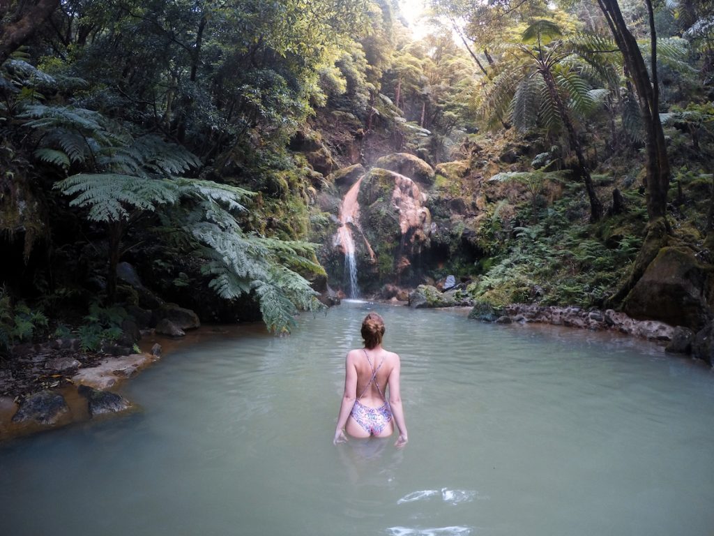 a woman in a bathing suit stairing at the waterfall in front of her in a thermal pool on a sao miguel vacation