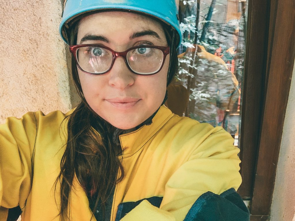 Allison Green, a white woman with. blue eyes, is wearing a yellow jumpsuit, red glasses, and a blue hard hat. She is giving an apprehensive smile.