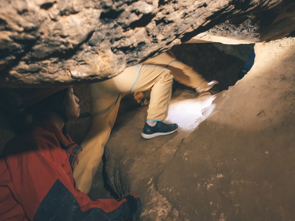 A person in a red hard hat with headlamp and red jumpsuit looking at someone in a yellow jumpsuit scrambling through a smaller crevice in the Budapest caves