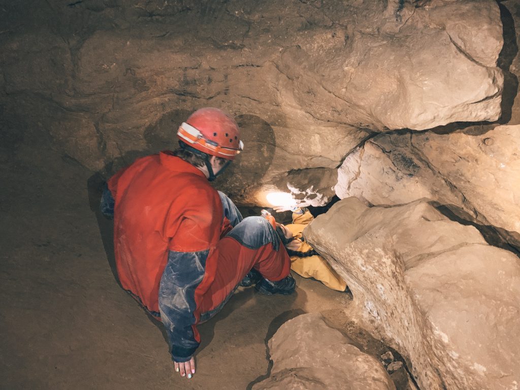 A person in an orange jumpsuit and orange hard hat going through a crevice in the Budapest caves with a person in yellow ahead of them.