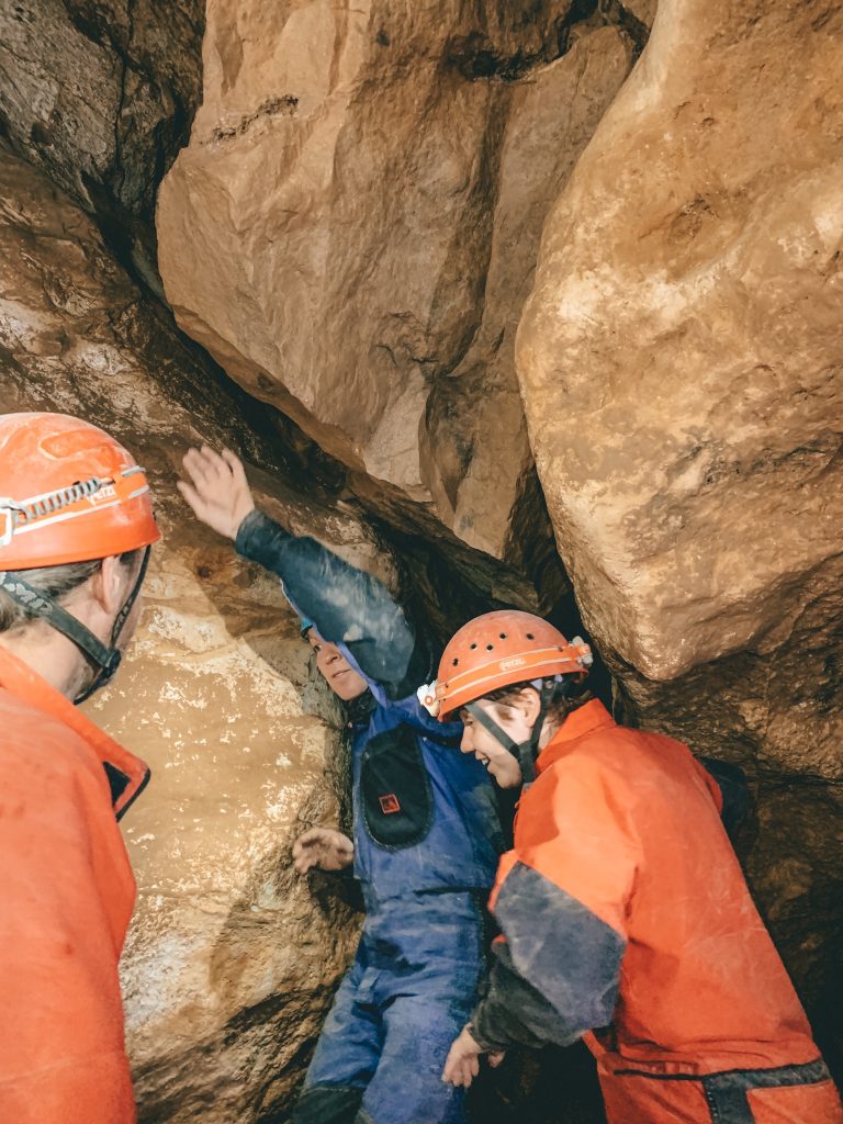 The guide in a blue jumpsuit and several people in orange jumpsuits and hard hats standing up while caving in Budapest