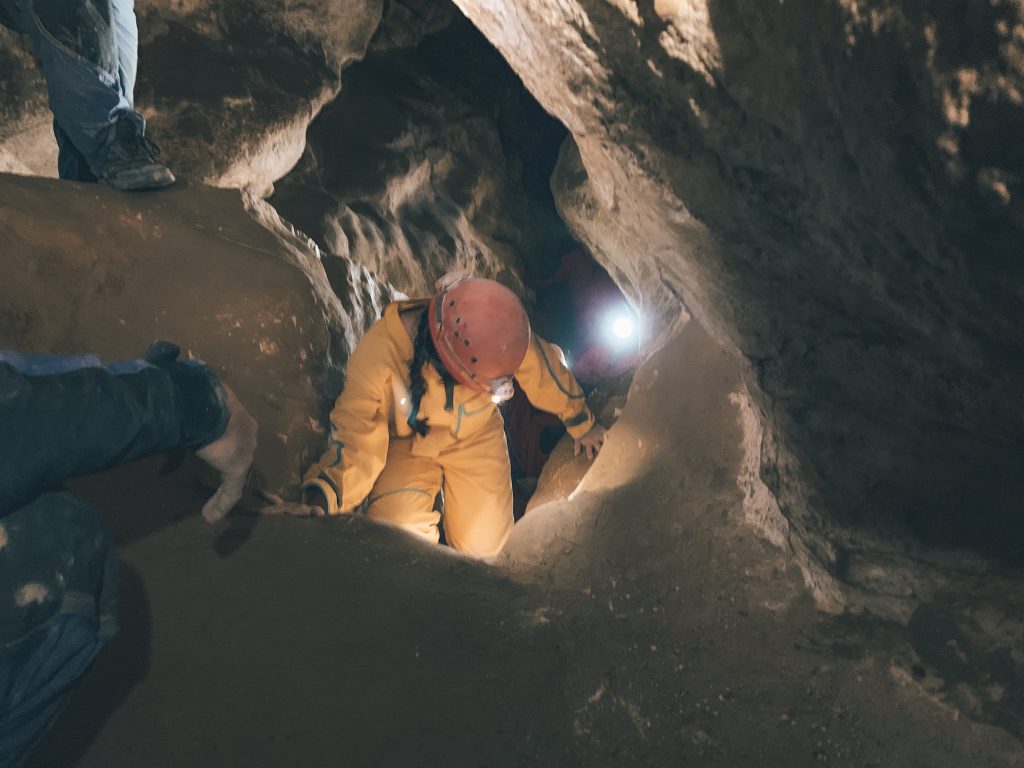 A person in a red hard hat with headlamp and yellow jumpsuit climbing up rocks on a Budapest cave tour, with the guide at the top to help