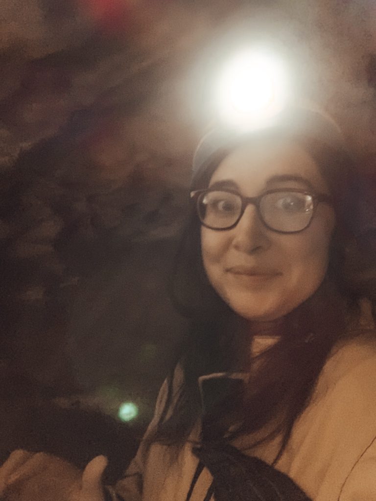 Allison Green smiling at the camera while wearing a yellow jumpsuit, hardhat, and headlamp while experiencing caving in Budapest.