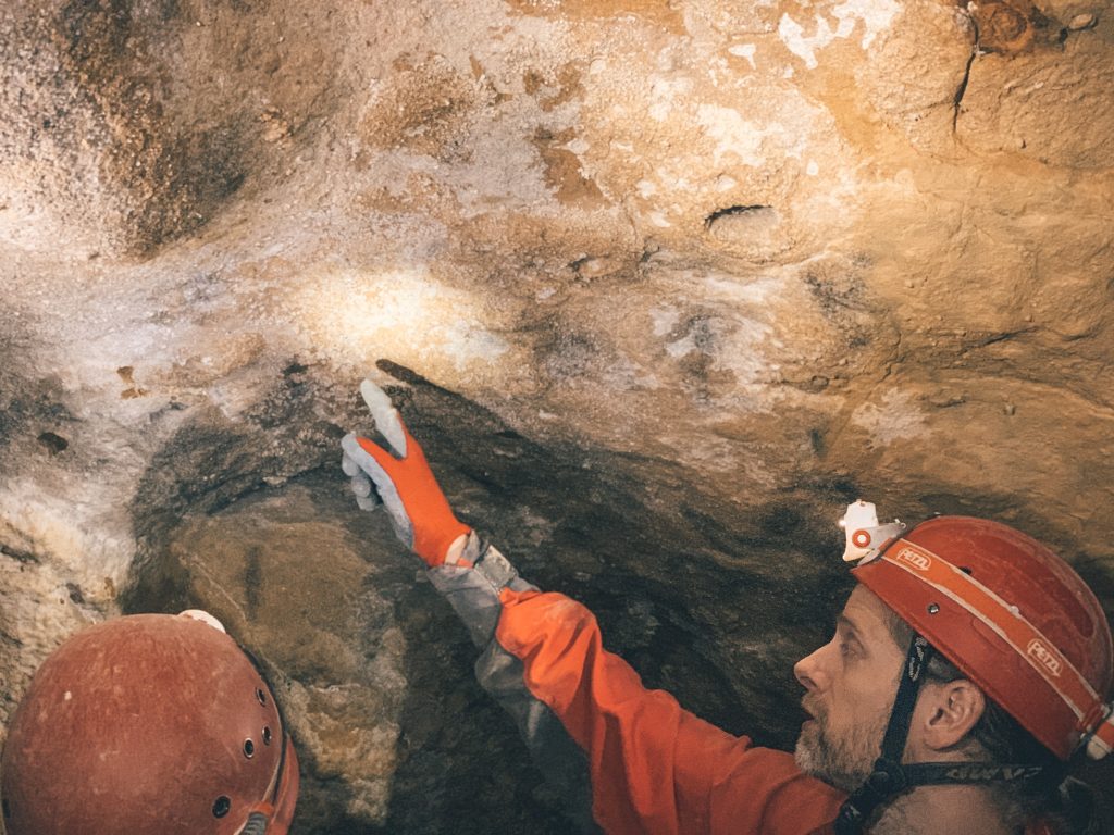 A man with a headlamp, hard hat, gloves, and caving jumpsuit pointing to a detail in the caves.