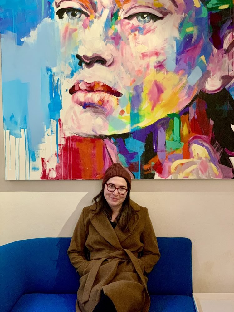 Allison in a peacoat, hat, glasses, sitting under a large piece of art