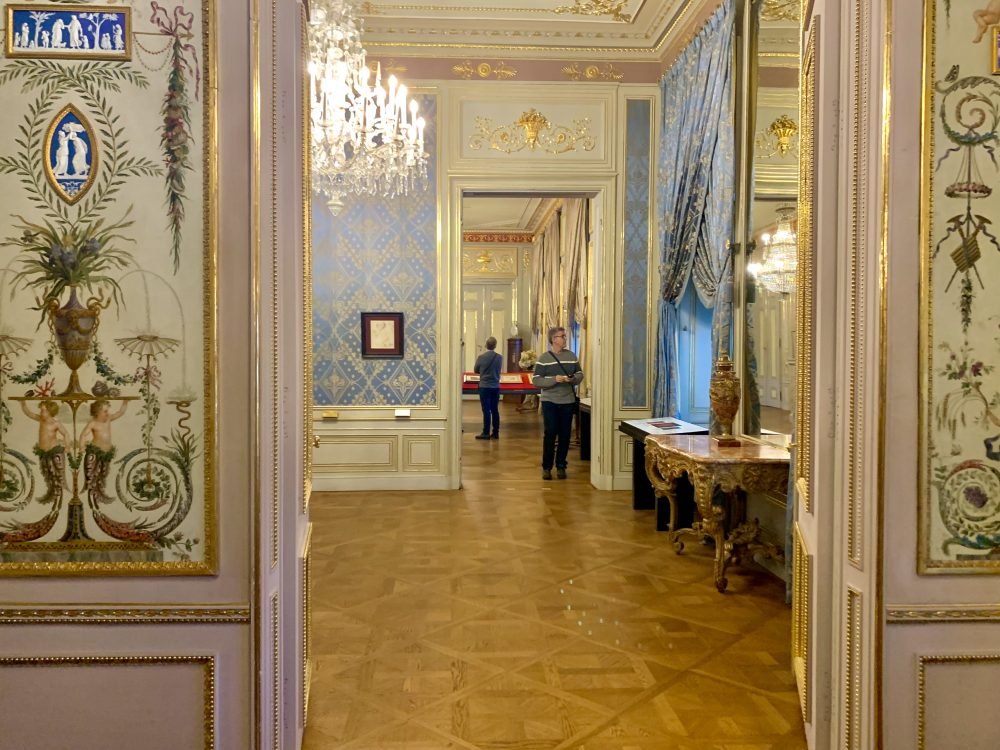 View at the Schonbrunn Palace of the interior with pastel, ornate wallpaper, and fancy home furnishings and people admiring the displays