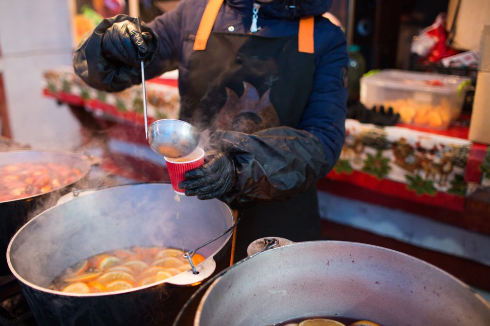 A person in their winter gear including cloves and a jacket and hood, pouring some hot mulled wine into a plastic cup with a ladel, steaming wine