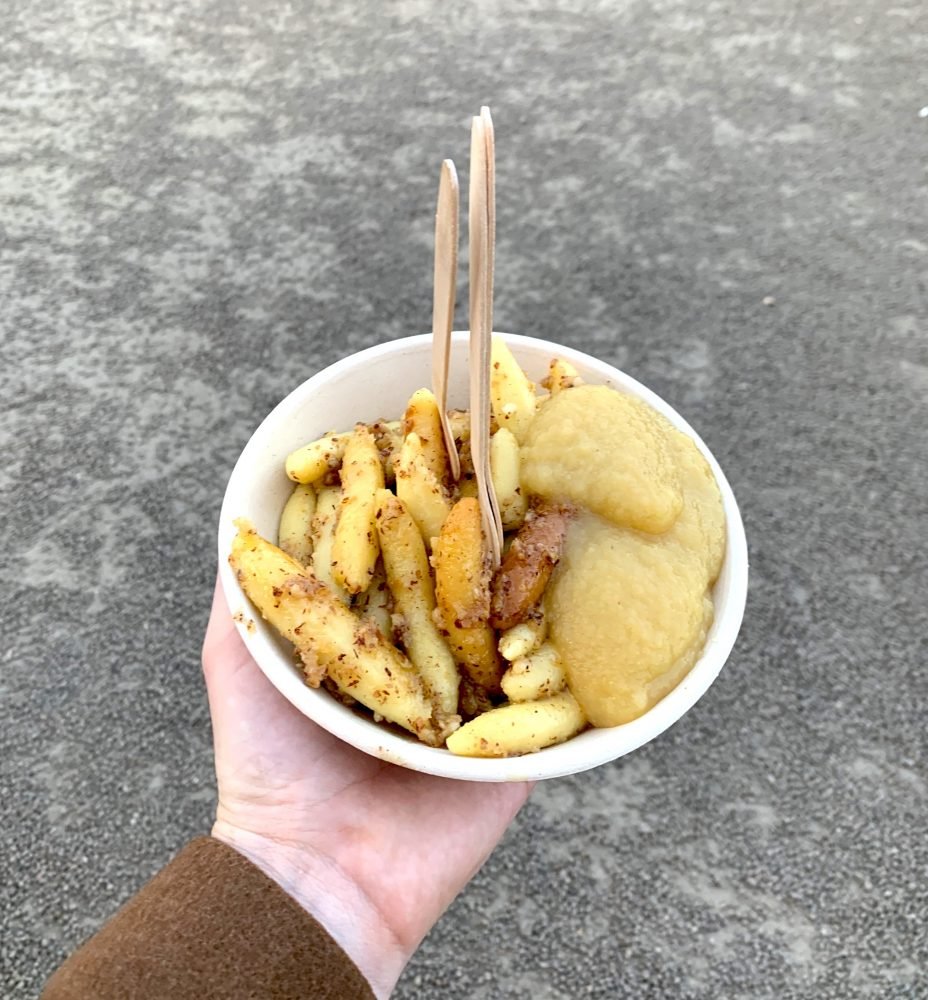 Allison's hand holding a thing of cheese spaetzel (dumplings) while wearing a winter jacket in Vienna