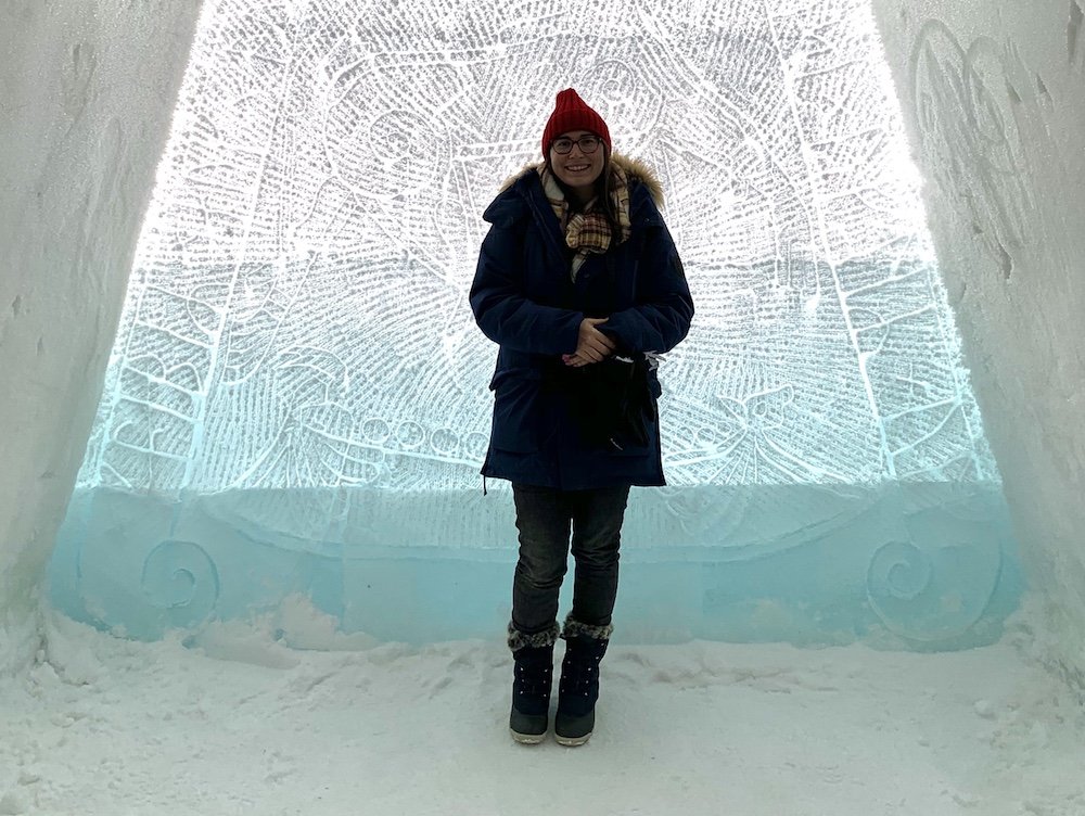 Allison wearing a red hat and blue jacket and snow boots and smiling in an ice hotel